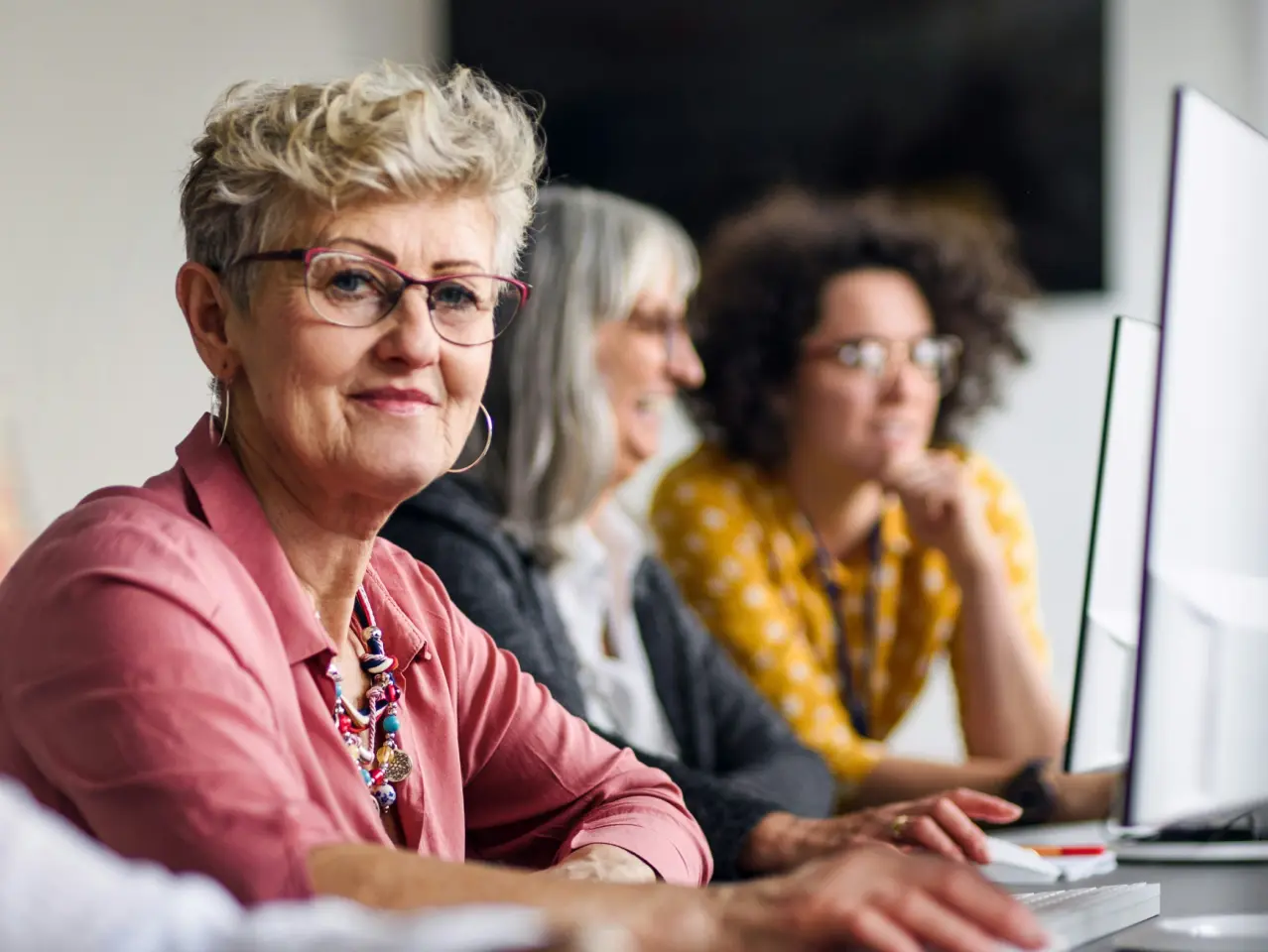 Smiling senior woman with glasses in pink shirt sitting at a desk with two other women working on computers.