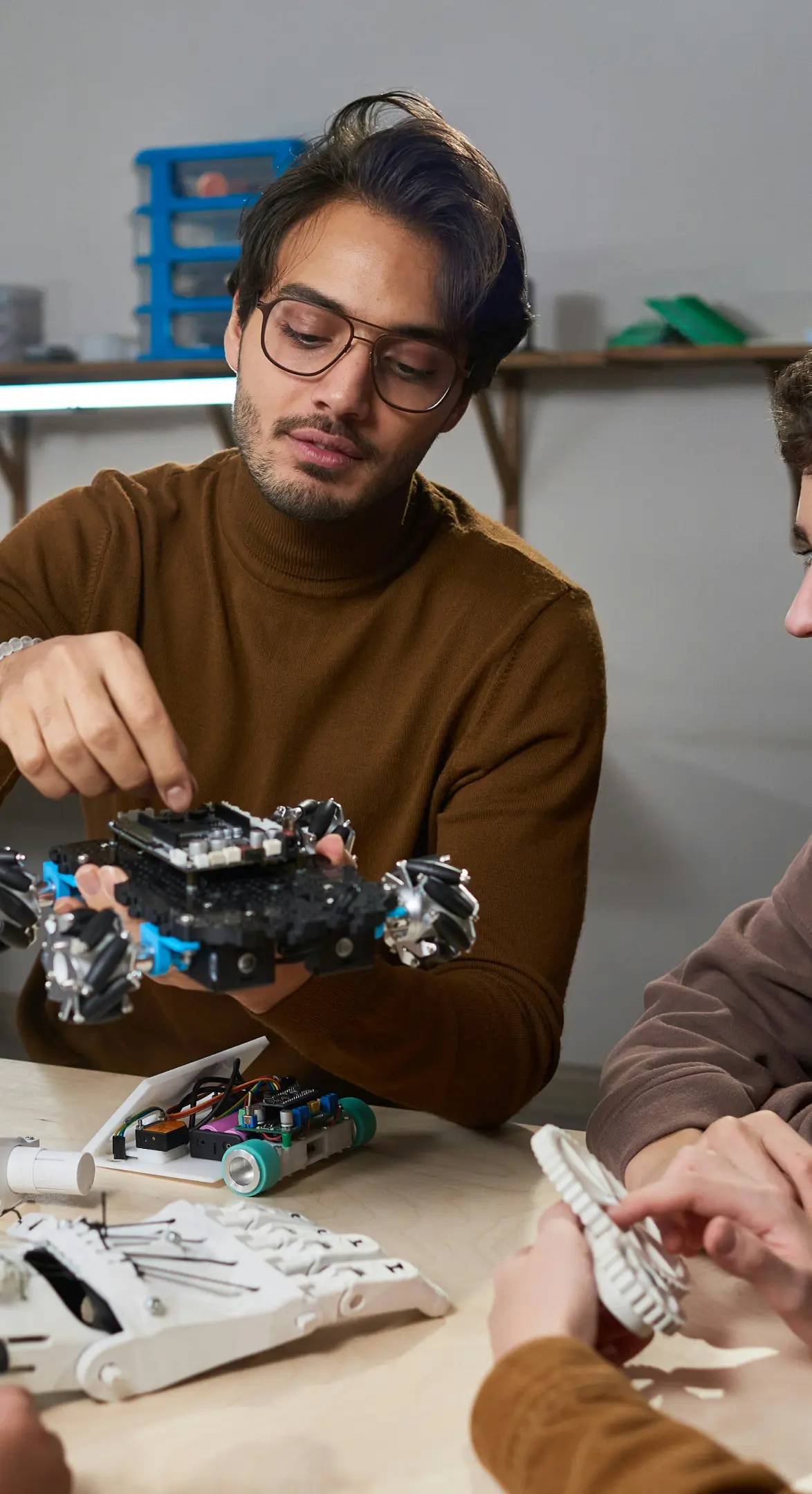 Man with glasses demonstrating a robotic device with complex wheels to two other people at a table containing electronic parts and a robotic hand.