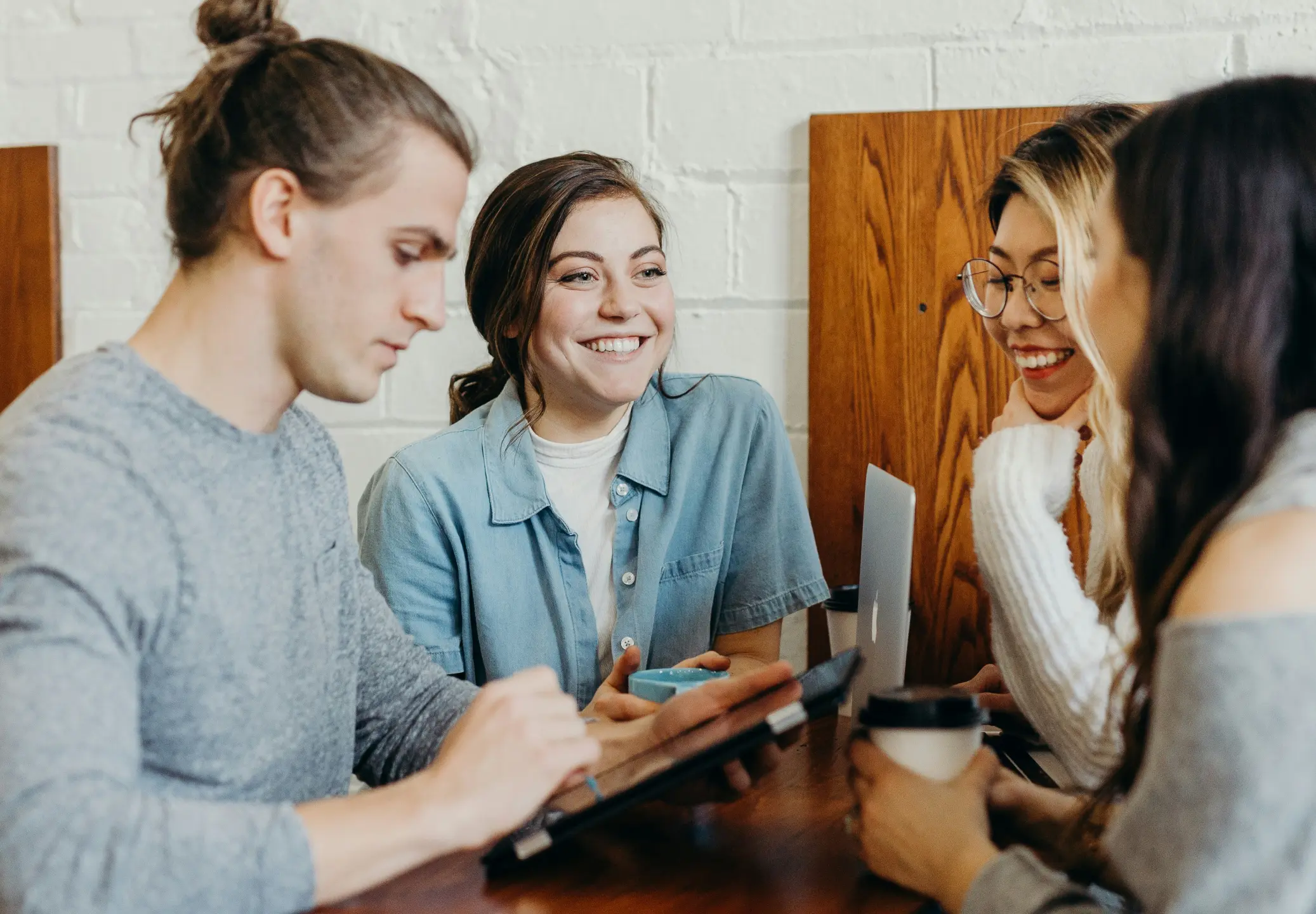 Four young adults sitting at a table, engaged in conversation and smiling, with two using laptops and two holding drinks.
