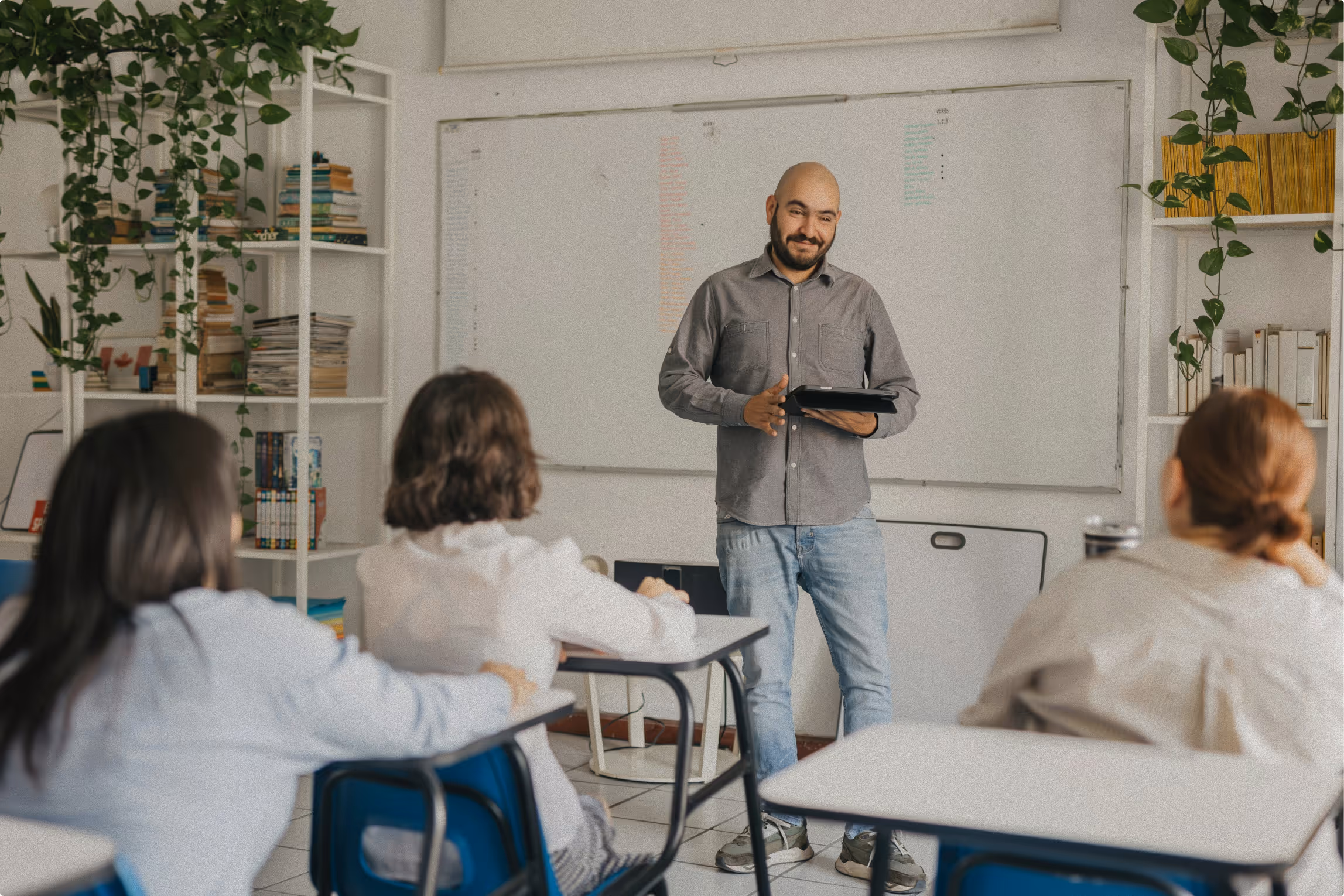 Man holding tablet and speaking to three seated students in a classroom with whiteboard and bookshelves.