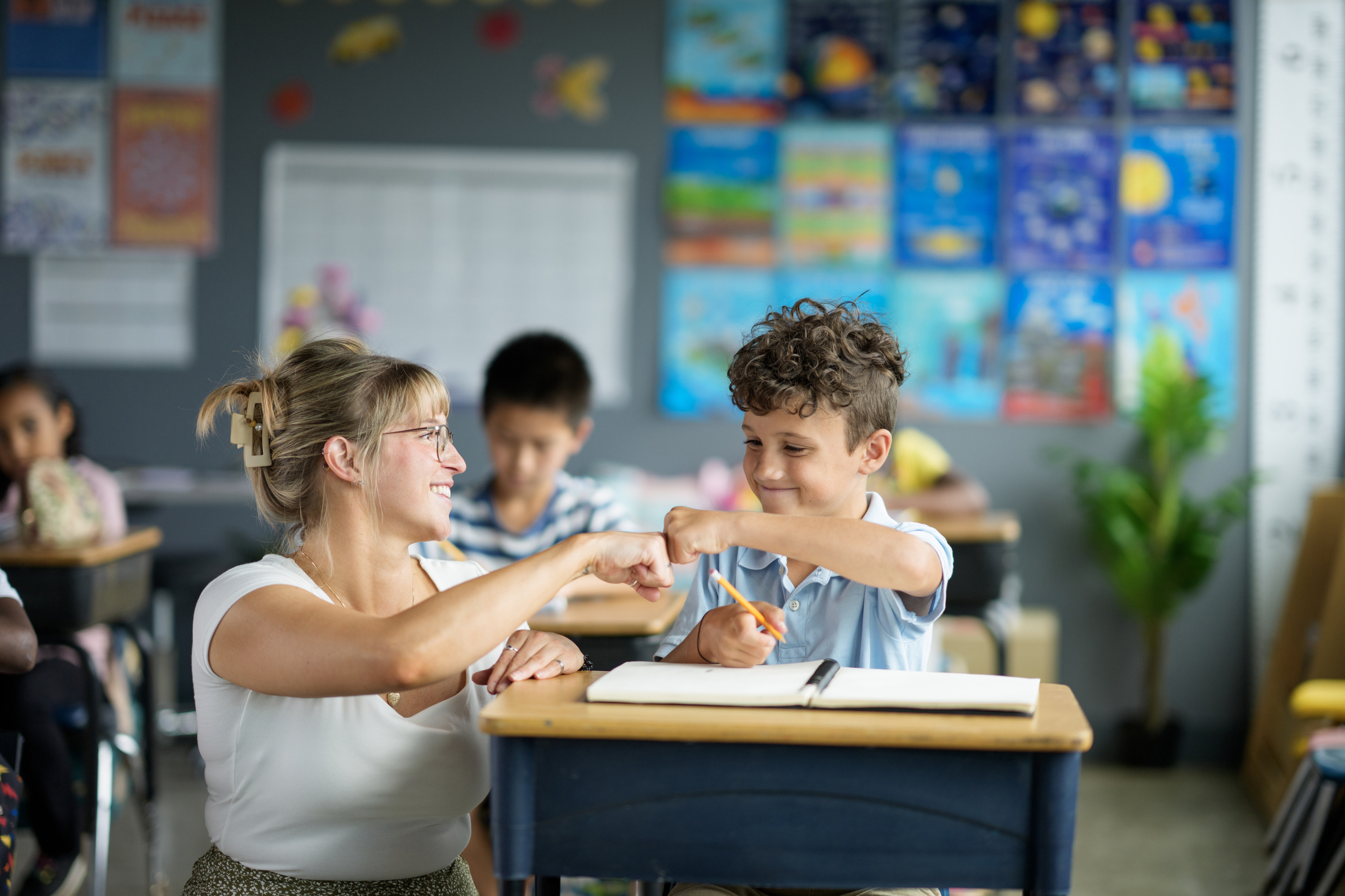 A teacher fist-bumps a smiling student at his desk in a classroom