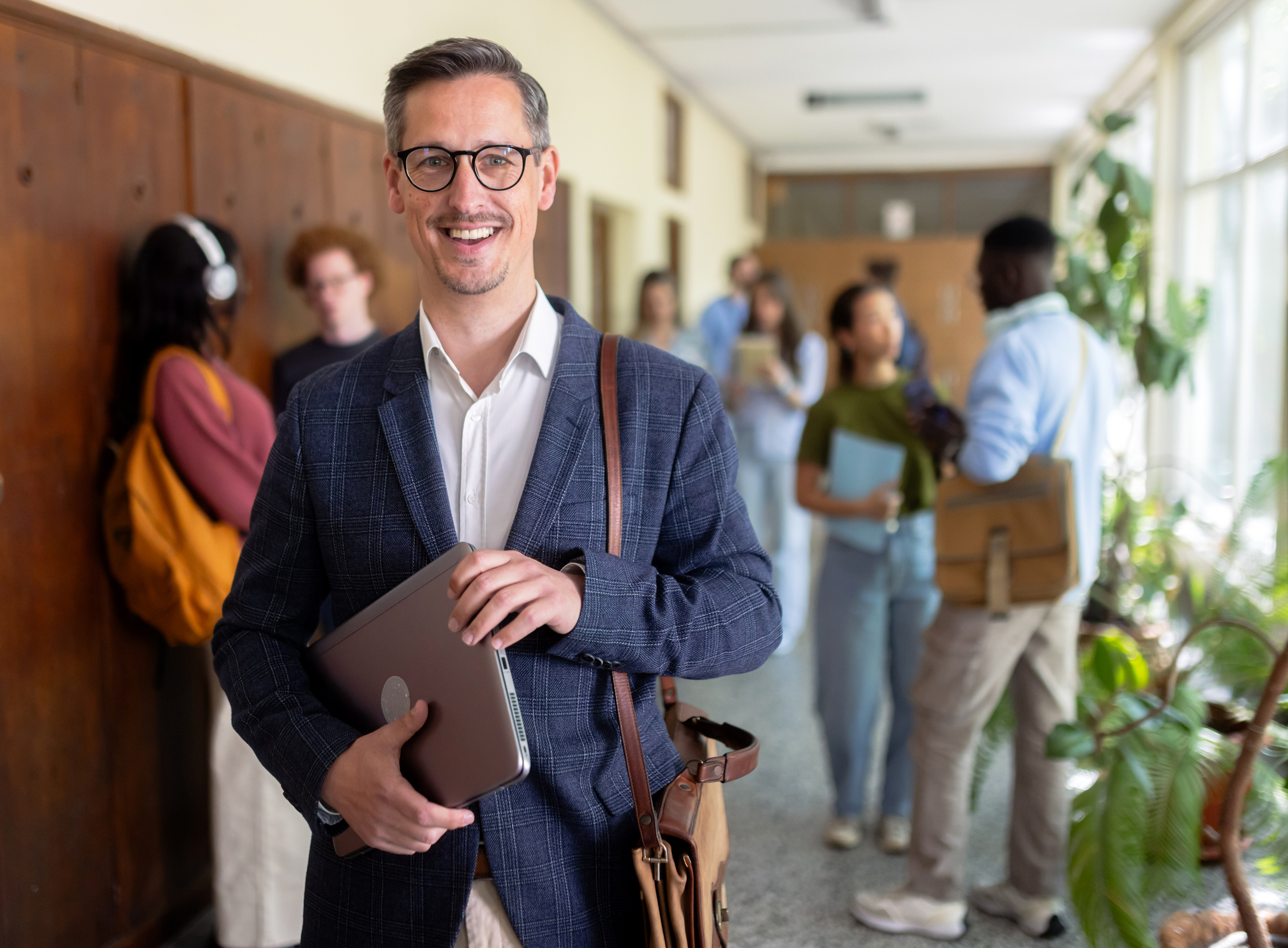 A smiling school principal holding a laptop in a busy school hallway