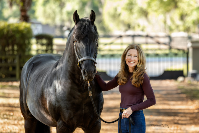 Smiling woman with long hair standing beside a large black horse holding its lead rope in a sunlit outdoor setting.