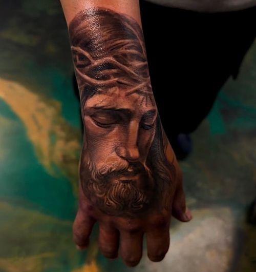 Realistic tattoo of Jesus Christ with crown of thorns on the back of a person's hand.
