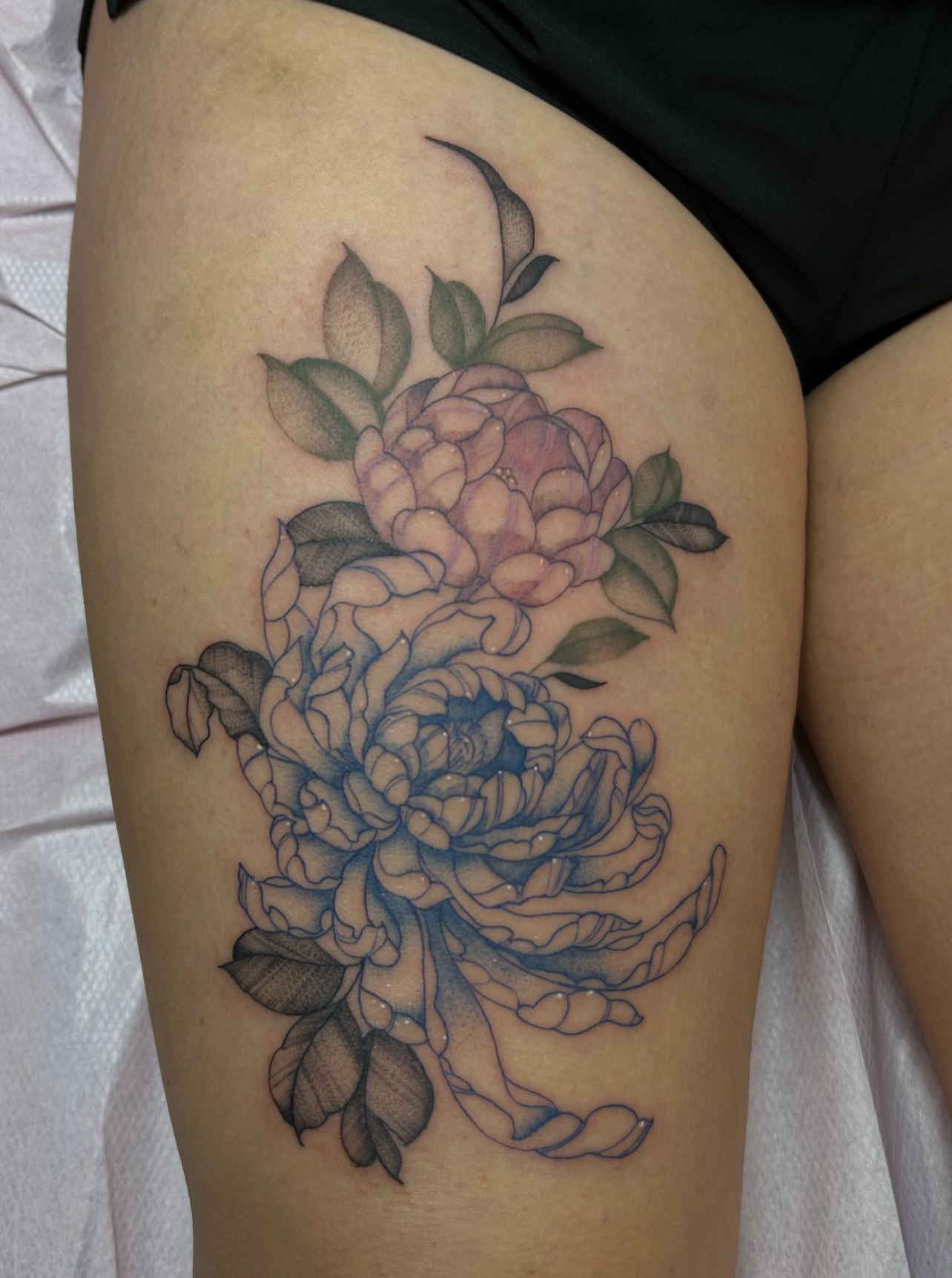 Thigh tattoo of two peony flowers with green leaves, one shaded in blue and the other in light pink.