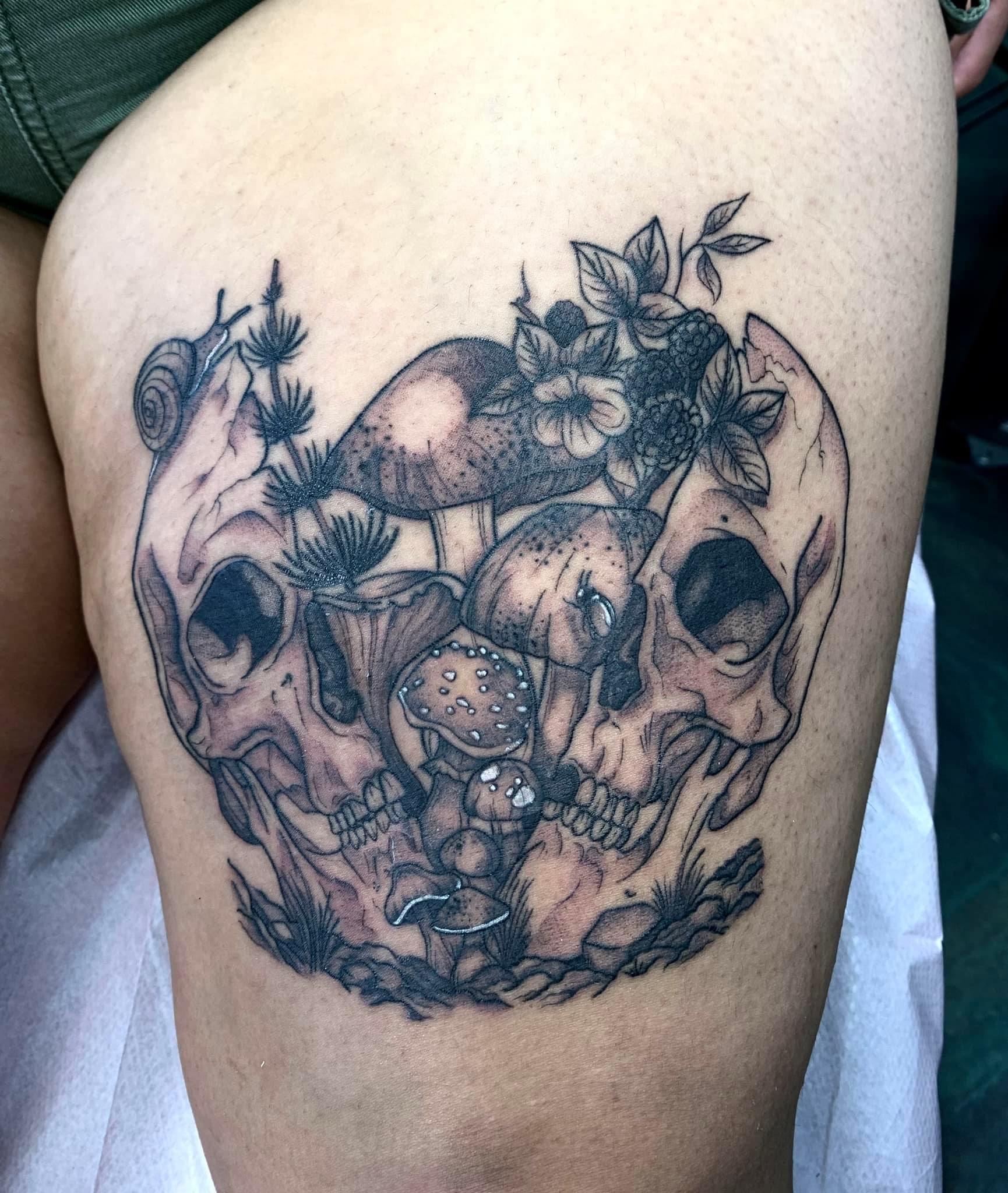Tattoo of two detailed skulls surrounded by various mushrooms, flowers, leaves, and a small snail.