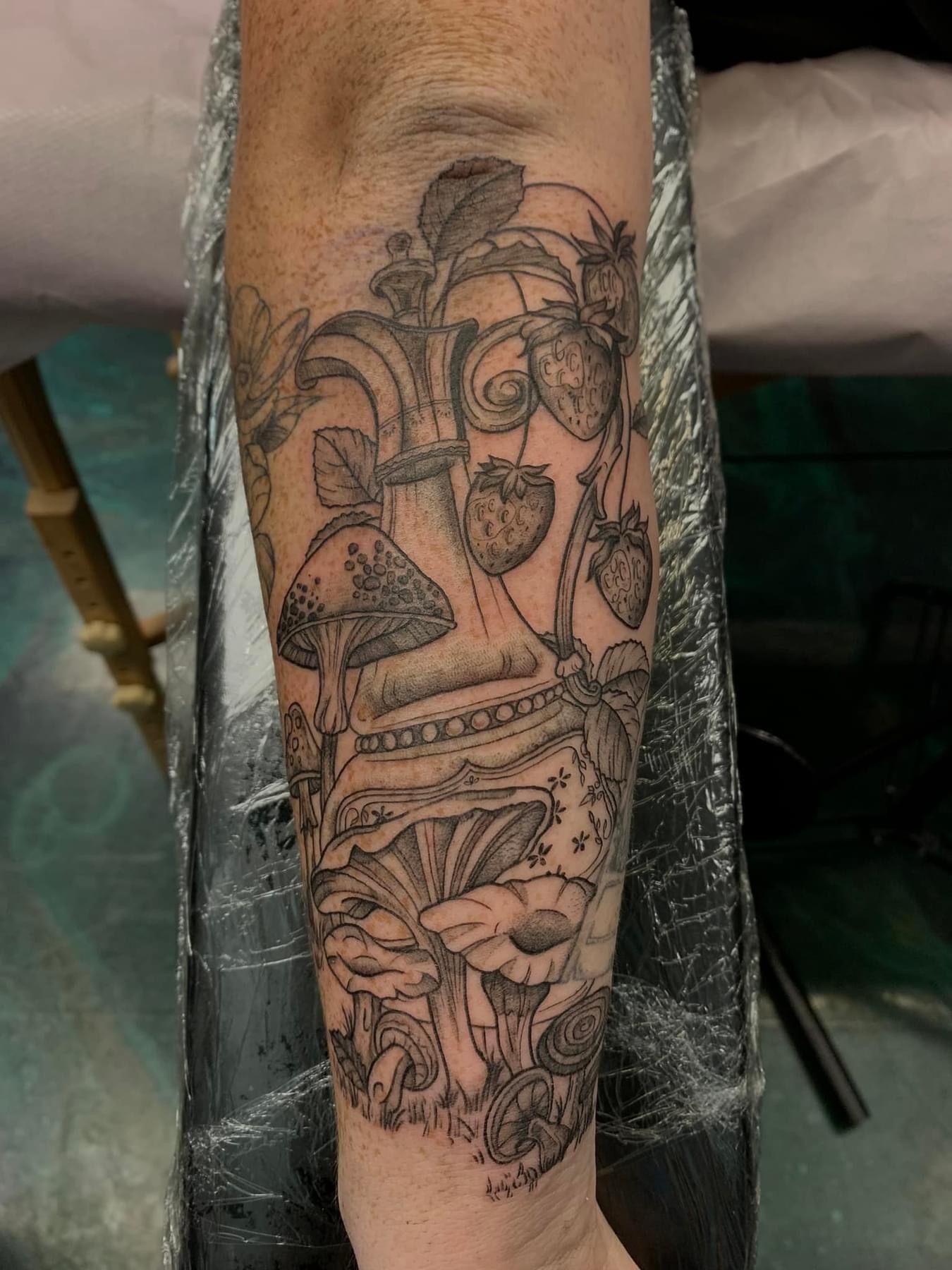 Fine line tattoo on forearm featuring a decorative bottle surrounded by strawberries, leaves, and various mushrooms.