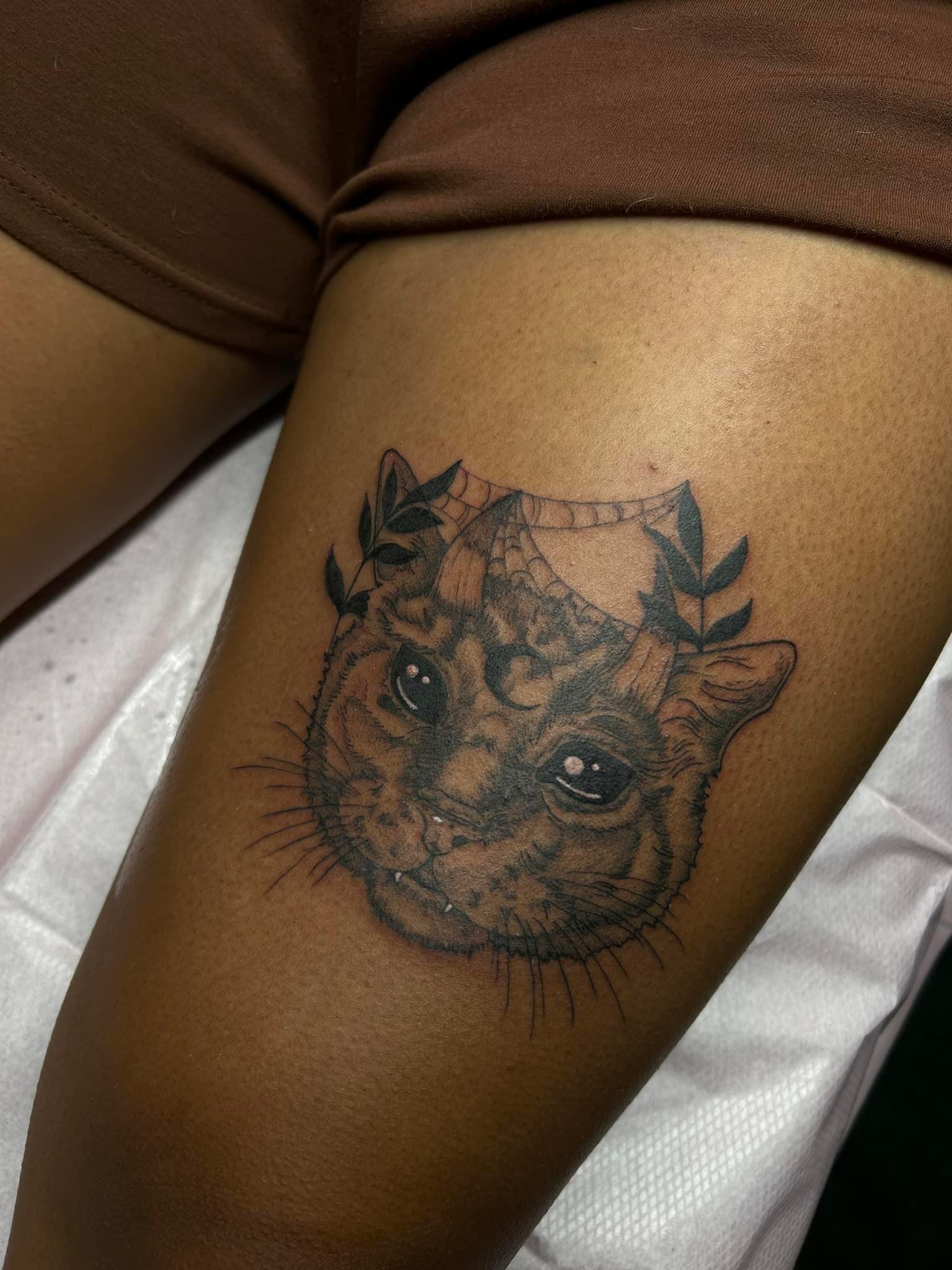 Tattoo of a cat head with a crescent moon on its forehead, surrounded by leaves and a spider web design.