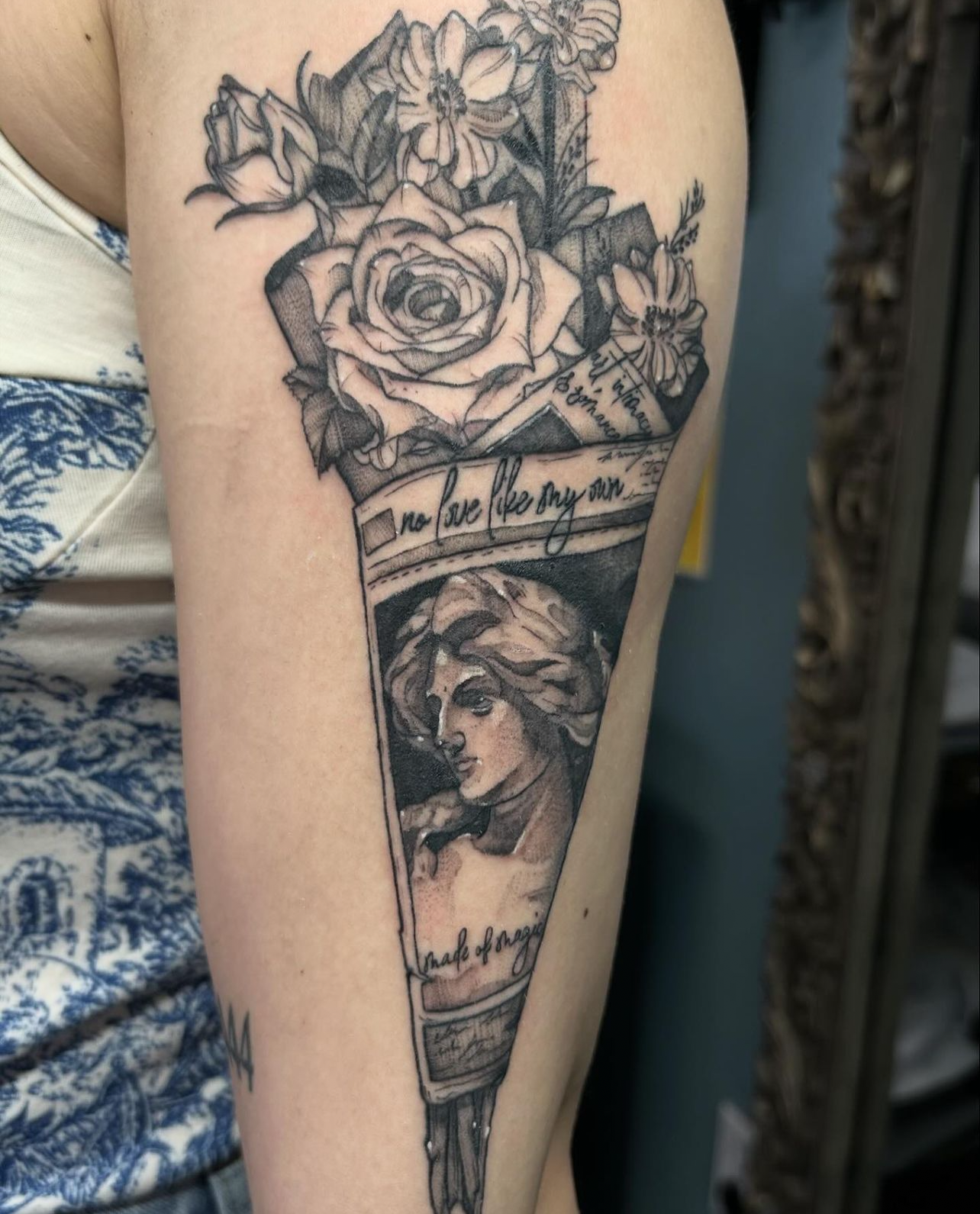 Fine line tattoo on upper arm featuring a classical statue bust inside a wrapped paper cone with roses and flowers.