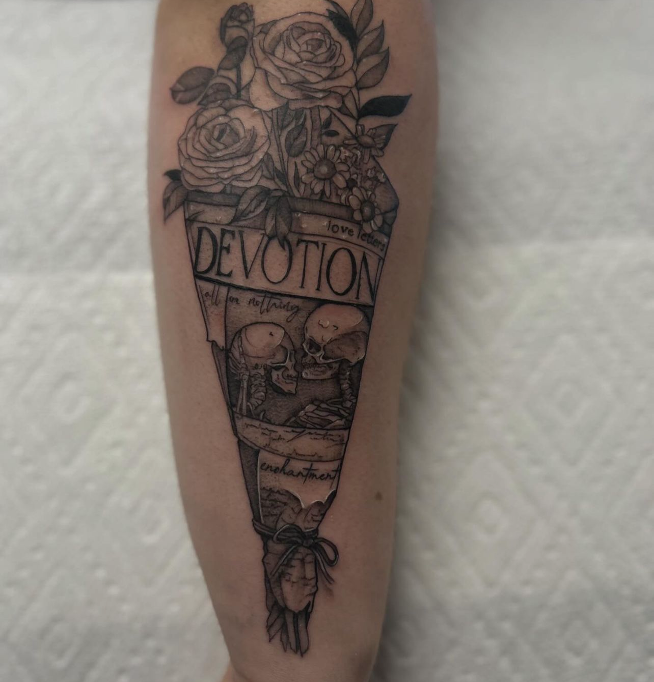 Fine line tattoo of a bouquet wrapped in paper with the word 'DEVOTION' and two skeletons facing each other.