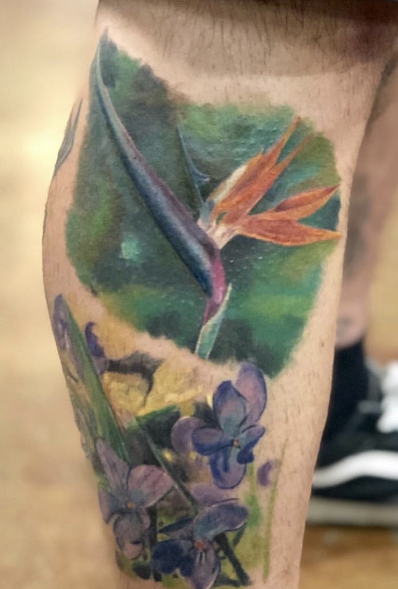 Tattoo of a bird of paradise flower with orange petals and purple flowers on a green leafy background.