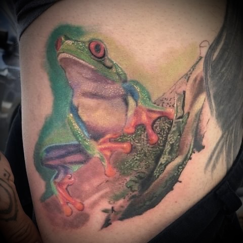 Colorful tattoo of a green frog with red eyes and orange toes sitting on a piece of bark.