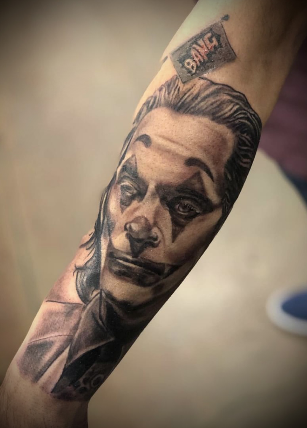 Black and gray tattoo of Joker with clown makeup and a 'BANG!' flag on a forearm.
