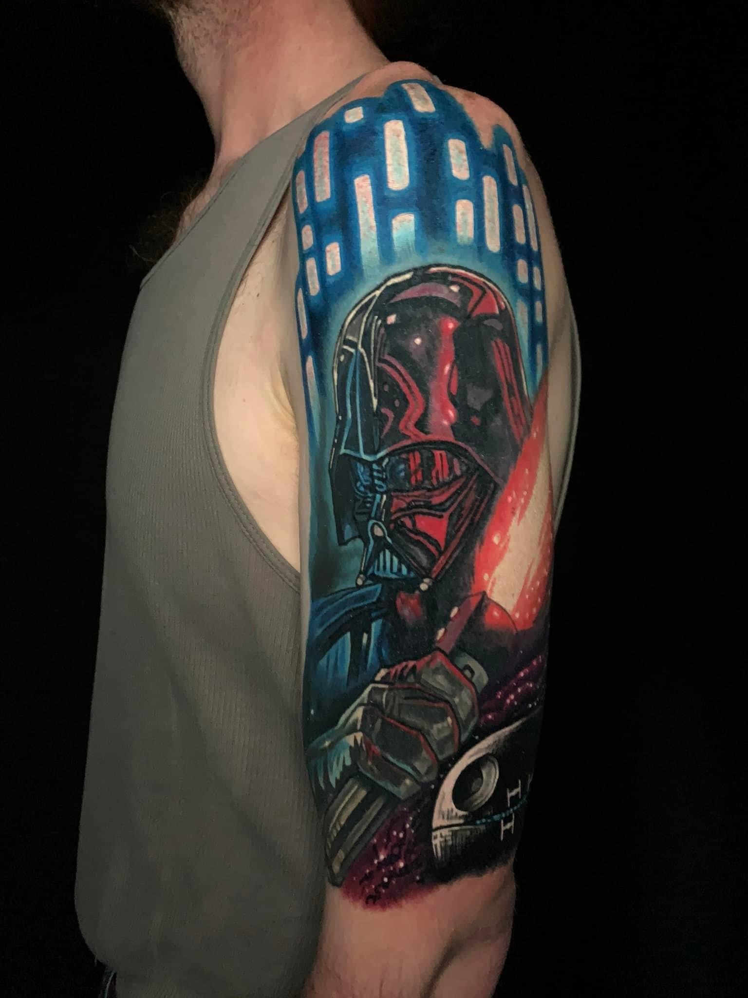Colorful tattoo of Darth Vader holding a red lightsaber with a Death Star in the background on a person's upper arm.
