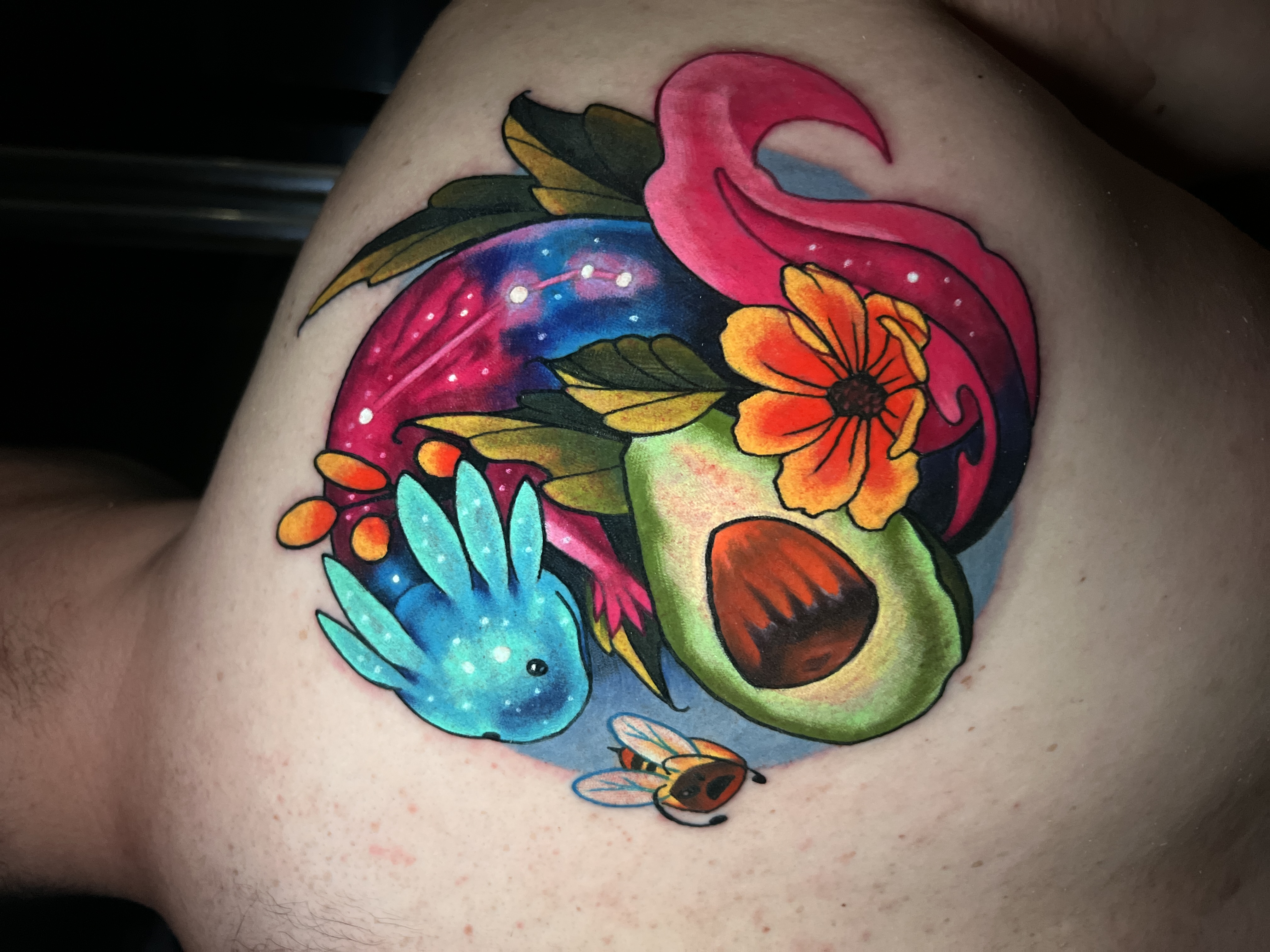 Colorful tattoo of a cosmic axolotl with orange gills, surrounded by an avocado half, orange flower, green leaves, and a small bee on a person's upper back.
