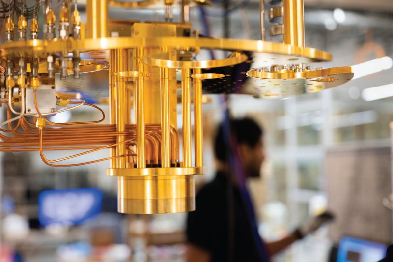 Close-up of a gold-colored quantum computer dilution refrigerator with copper wiring and a blurred person in the background.