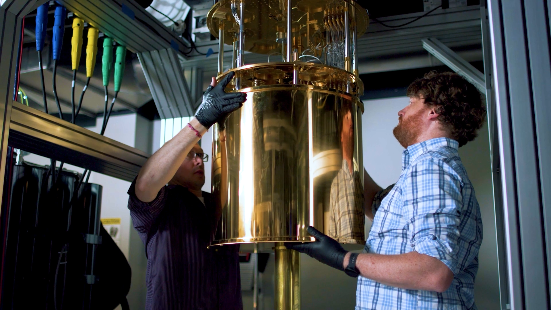 Two men wearing black gloves carefully handling a large, polished gold-colored cylindrical device in a laboratory setting.