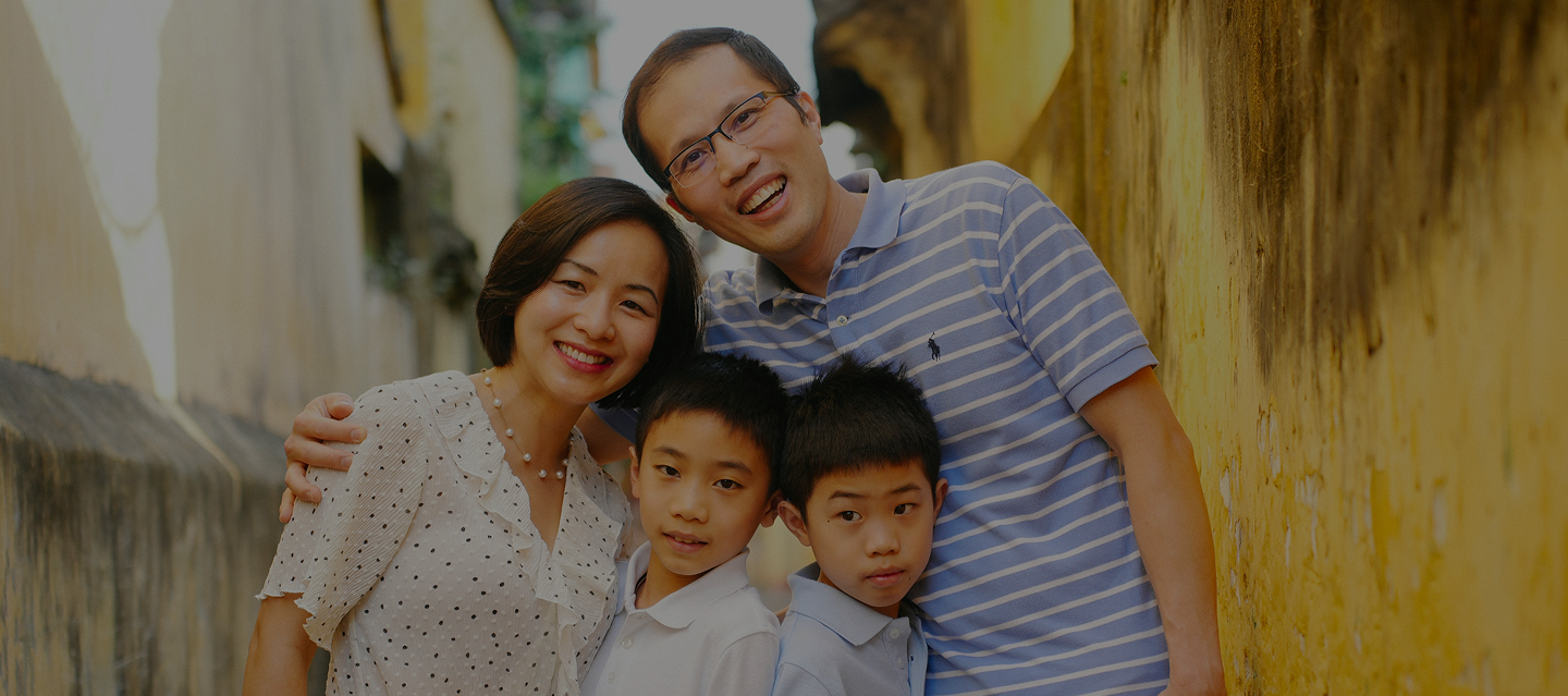 Smiling Asian family of four standing closely together in a narrow outdoor alley with yellow walls.