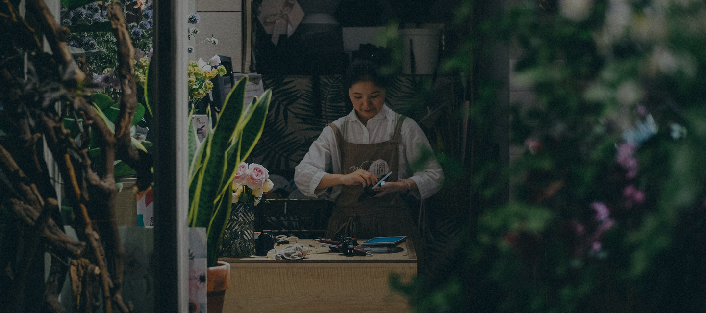 Florist in an apron arranging flowers and tools at a wooden workstation surrounded by plants and floral decorations.