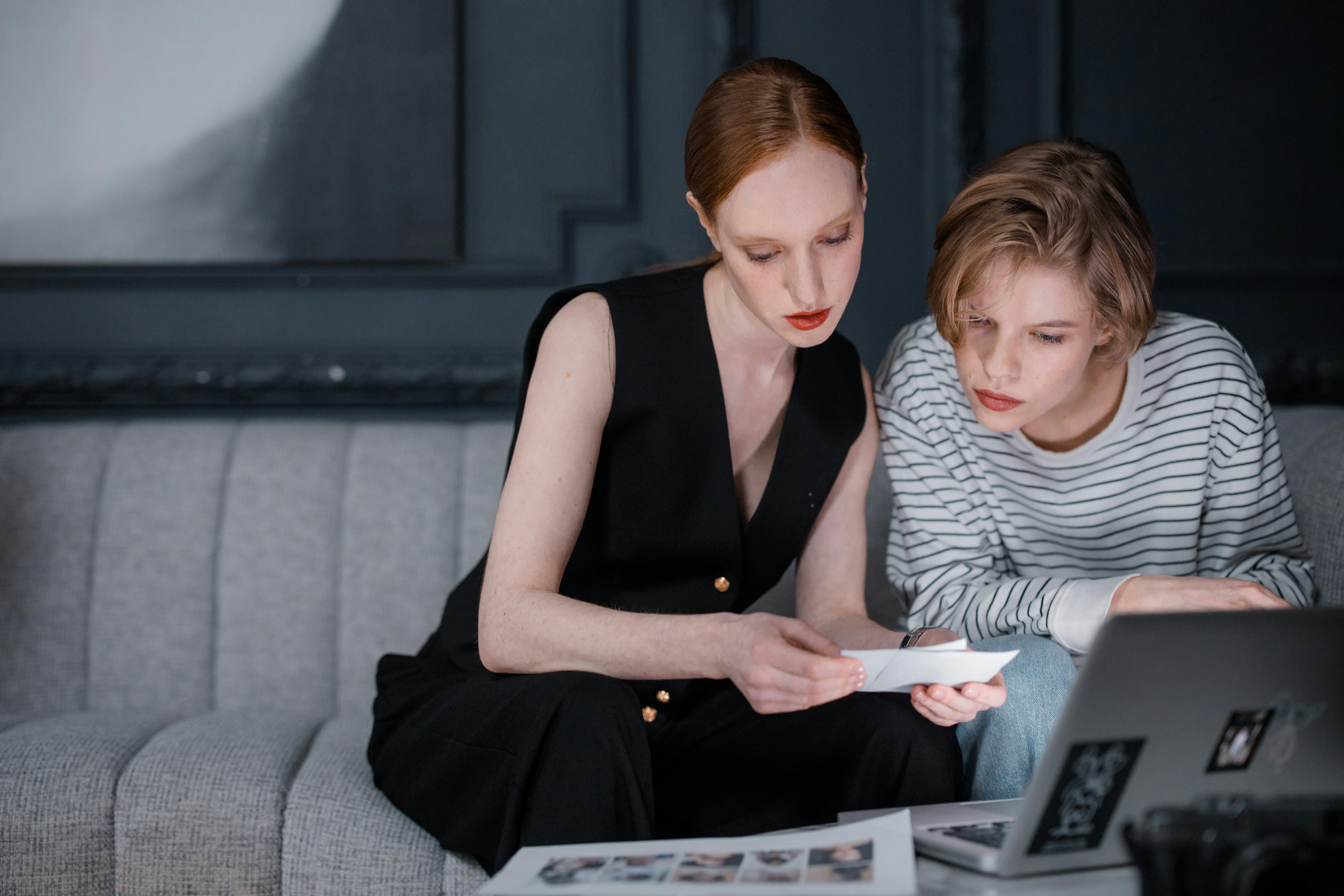 Two women sitting on a grey couch reviewing photos together, one holding a printed photo and the other looking at a laptop.