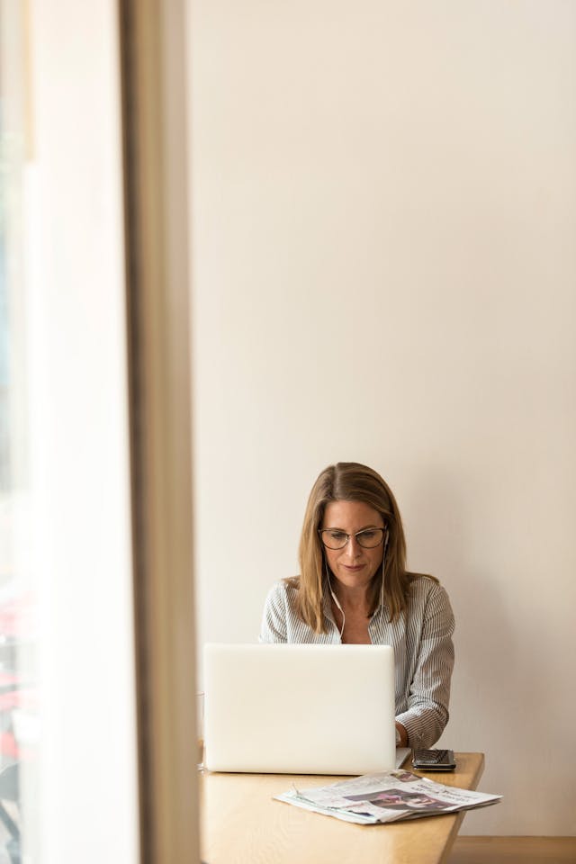 Woman with glasses sitting at a table working on a laptop with newspapers and a phone nearby.