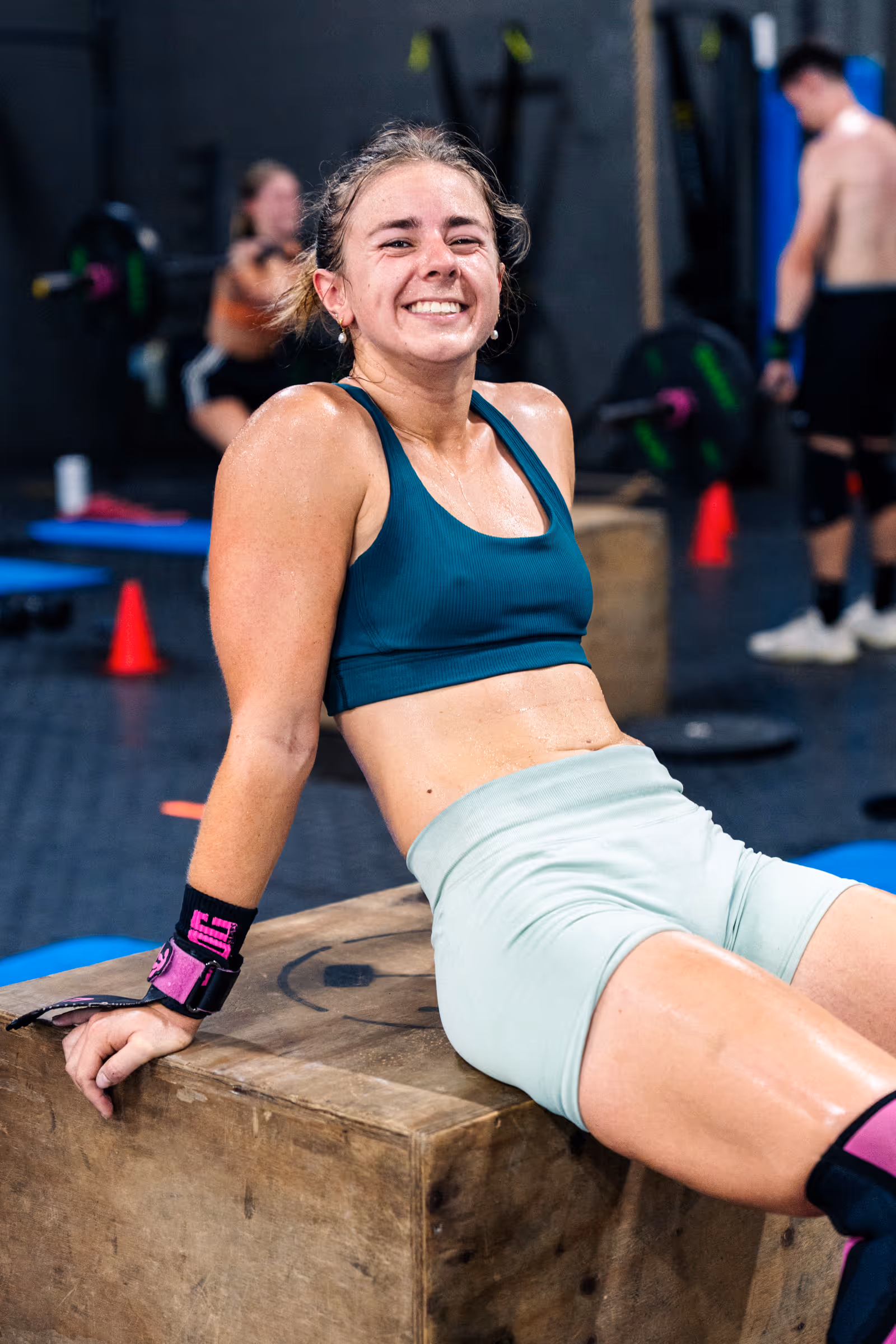 Smiling woman in workout clothes sitting on a wooden box in a gym after exercise.