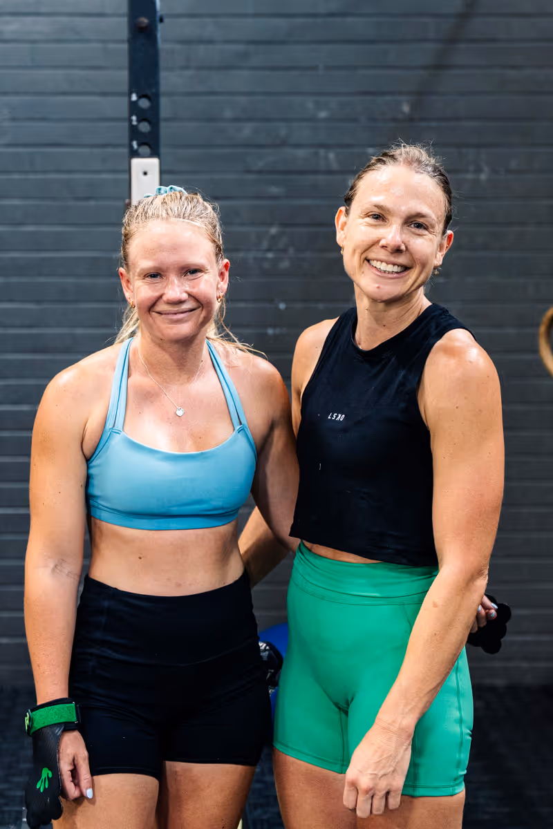 Two smiling women in workout clothes standing together in a gym with a gray wall background.