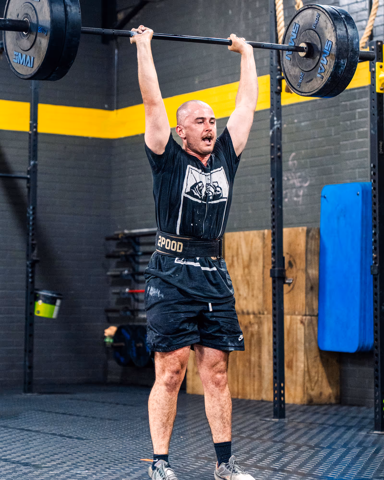 Man lifting a heavy barbell overhead in a gym wearing a lifting belt.