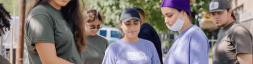 A diverse group of young adults standing outdoors, some wearing casual clothes and one wearing a face mask under their chin.