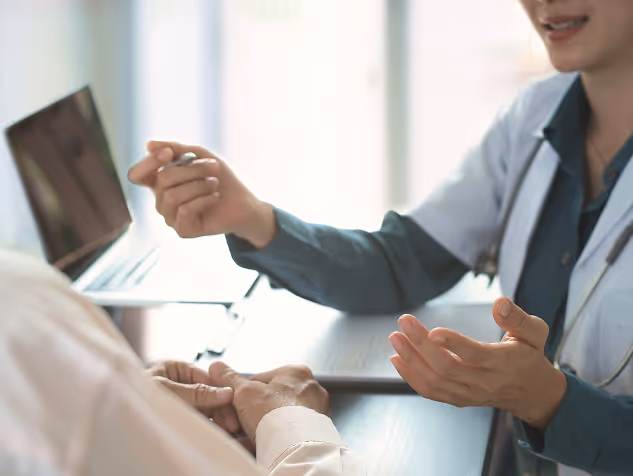 Doctor in a white coat discussing with a patient, gesturing with hands at a desk with a laptop.