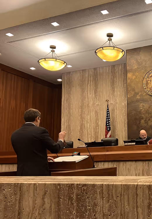 A man in a suit speaking at a podium in a formal courtroom setting with a judge seated to the right and an American flag behind them.