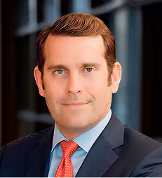 Portrait of a man in a dark blue suit, white shirt, and red tie with a blurred office background.