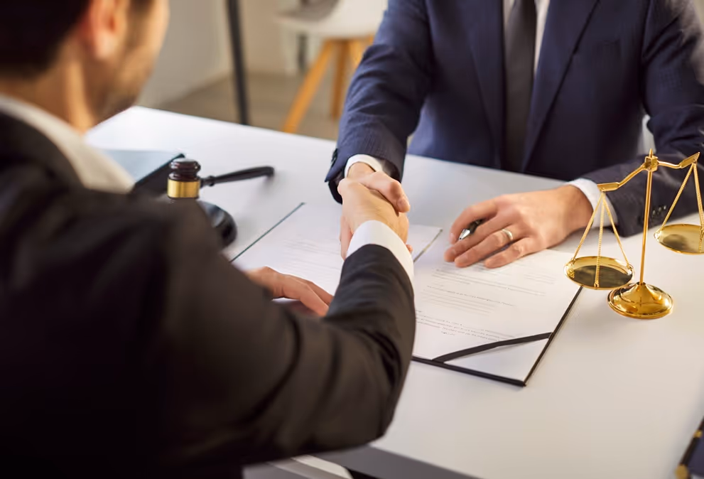 Two men in suits shaking hands across a desk with legal documents, a gavel, and balance scales.