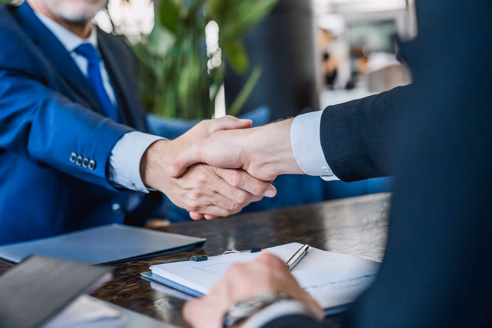 Two businessmen shaking hands over a desk with documents and a pen.