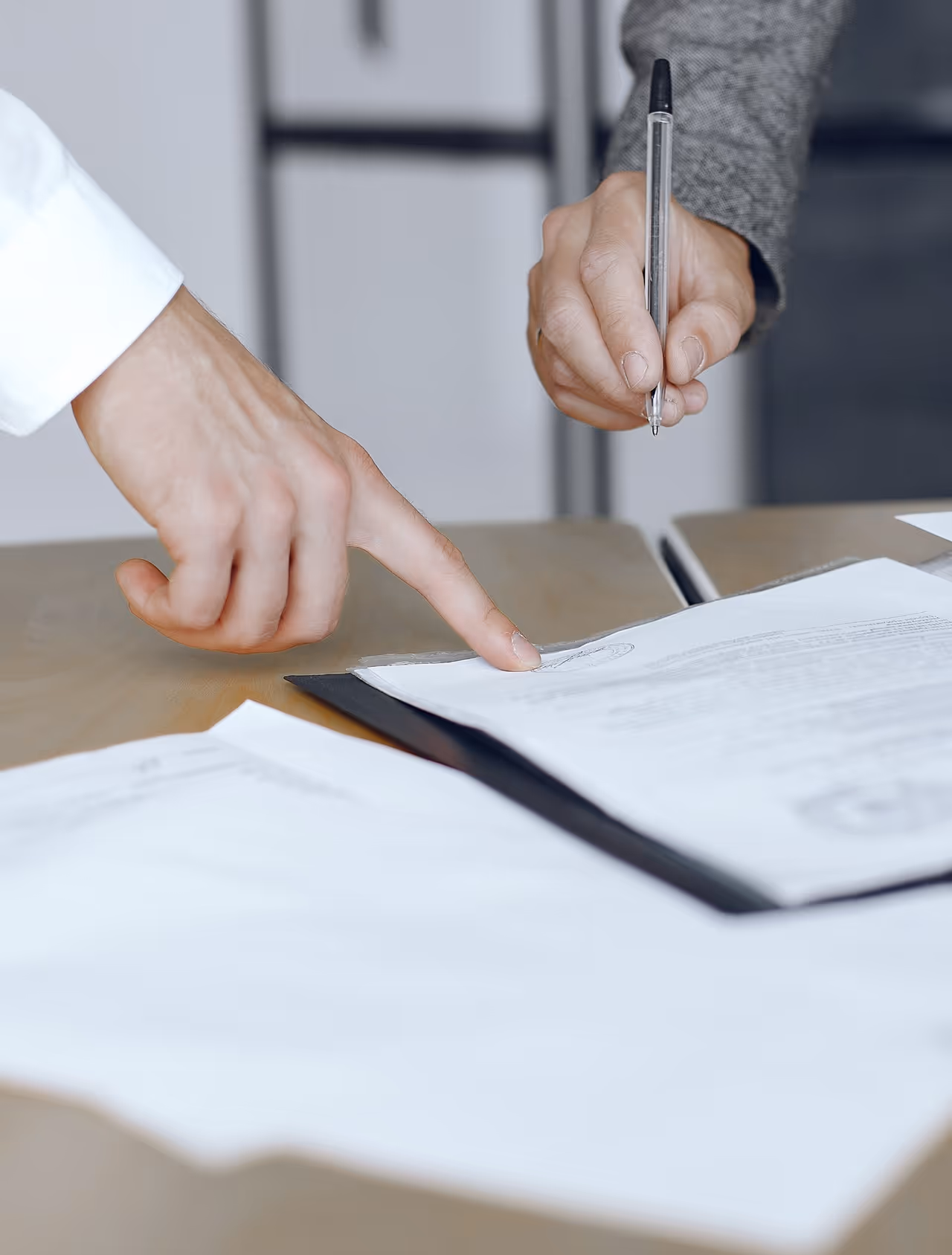 A person pointing at a document on a table while another hand holds a pen above it.