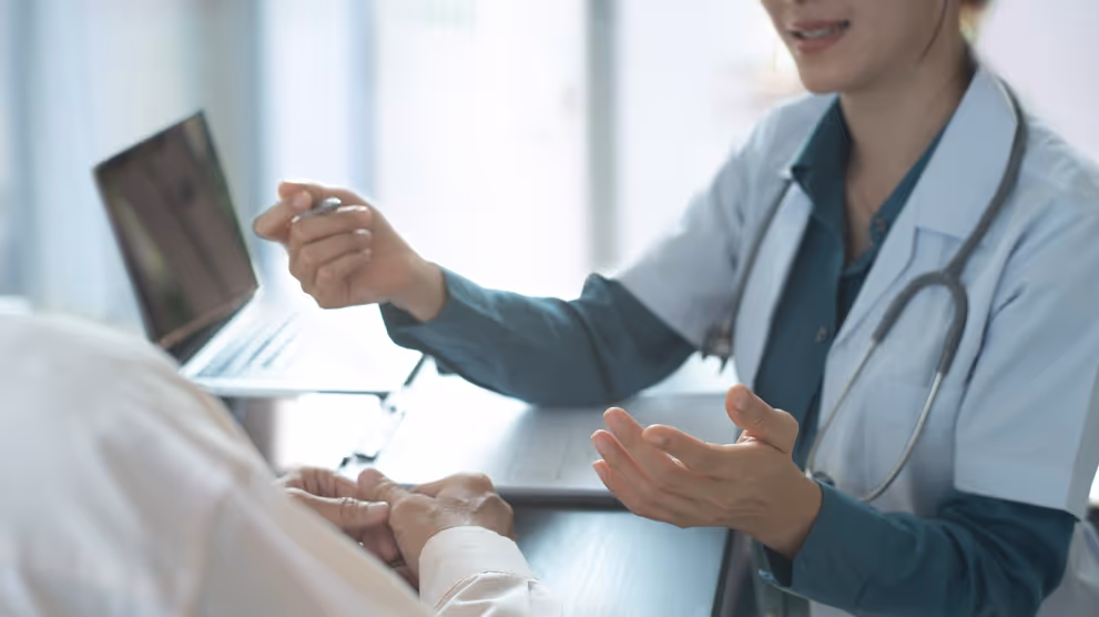 Doctor in white coat with stethoscope talking and gesturing with hands to a patient across a desk with a laptop.