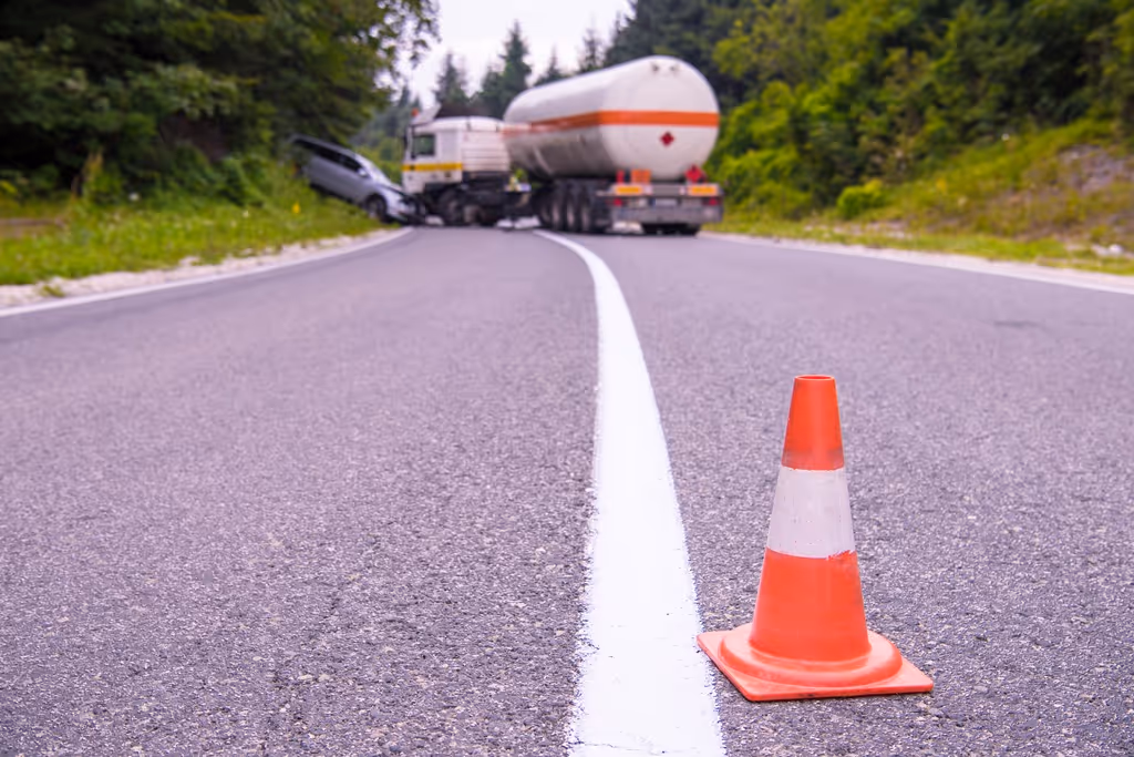 Orange traffic cone on a road with a tanker truck and a crashed car in the background near a forested area.