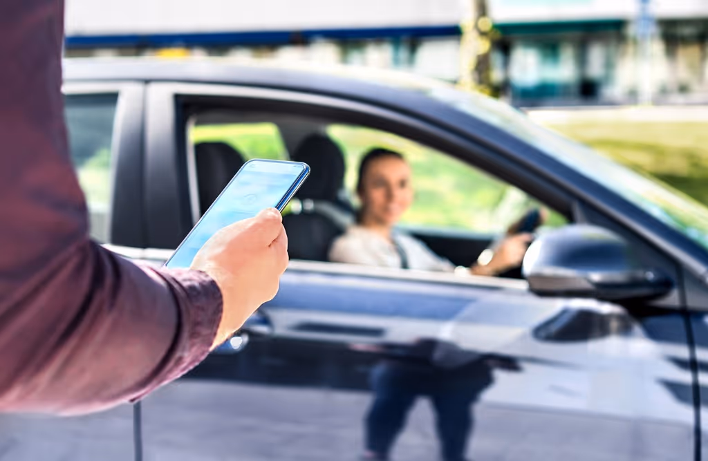 Person holding a smartphone near a car with a woman sitting inside the driver's seat.