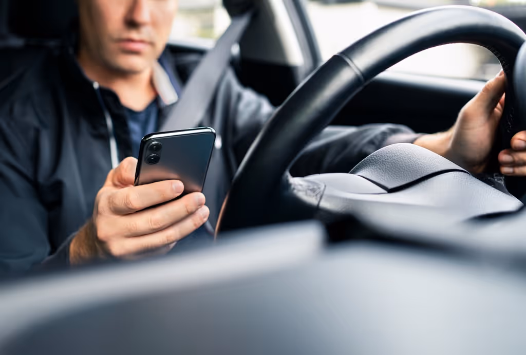 Driver wearing a seatbelt looking at a smartphone while holding the steering wheel with one hand.