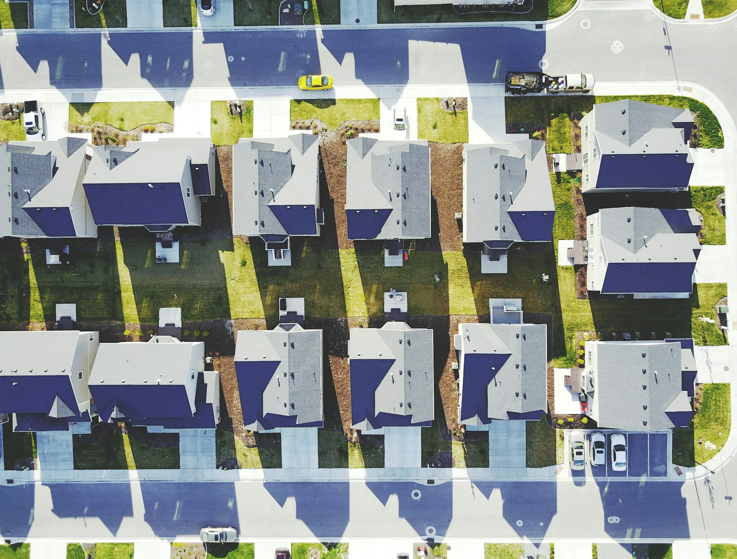 Aerial view of a residential neighborhood with rows of single-family houses, green lawns, driveways, and parked cars casting long shadows.