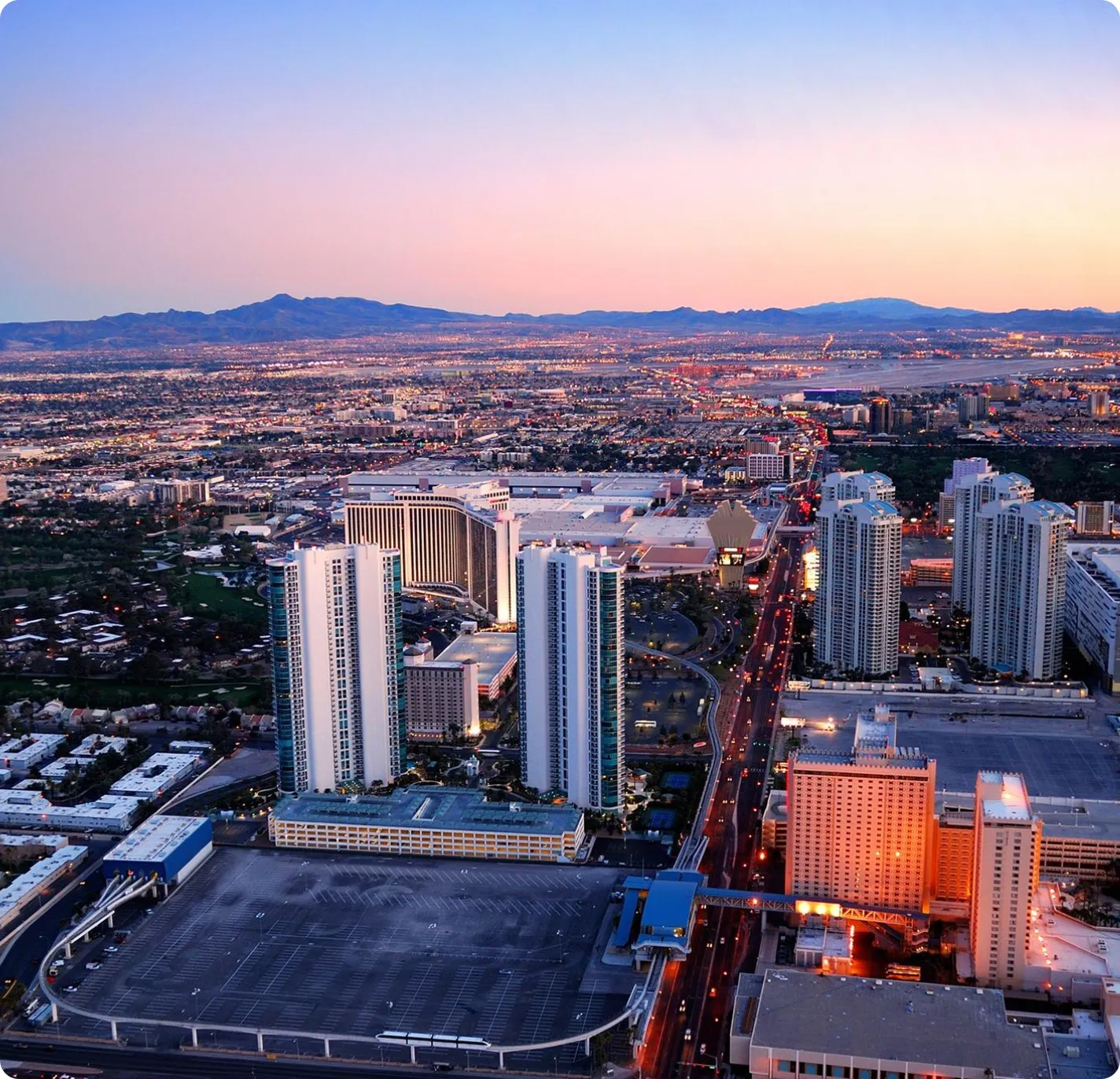 Aerial view of a cityscape at dusk with high-rise buildings, a large parking lot, and distant mountains under a pastel sky.