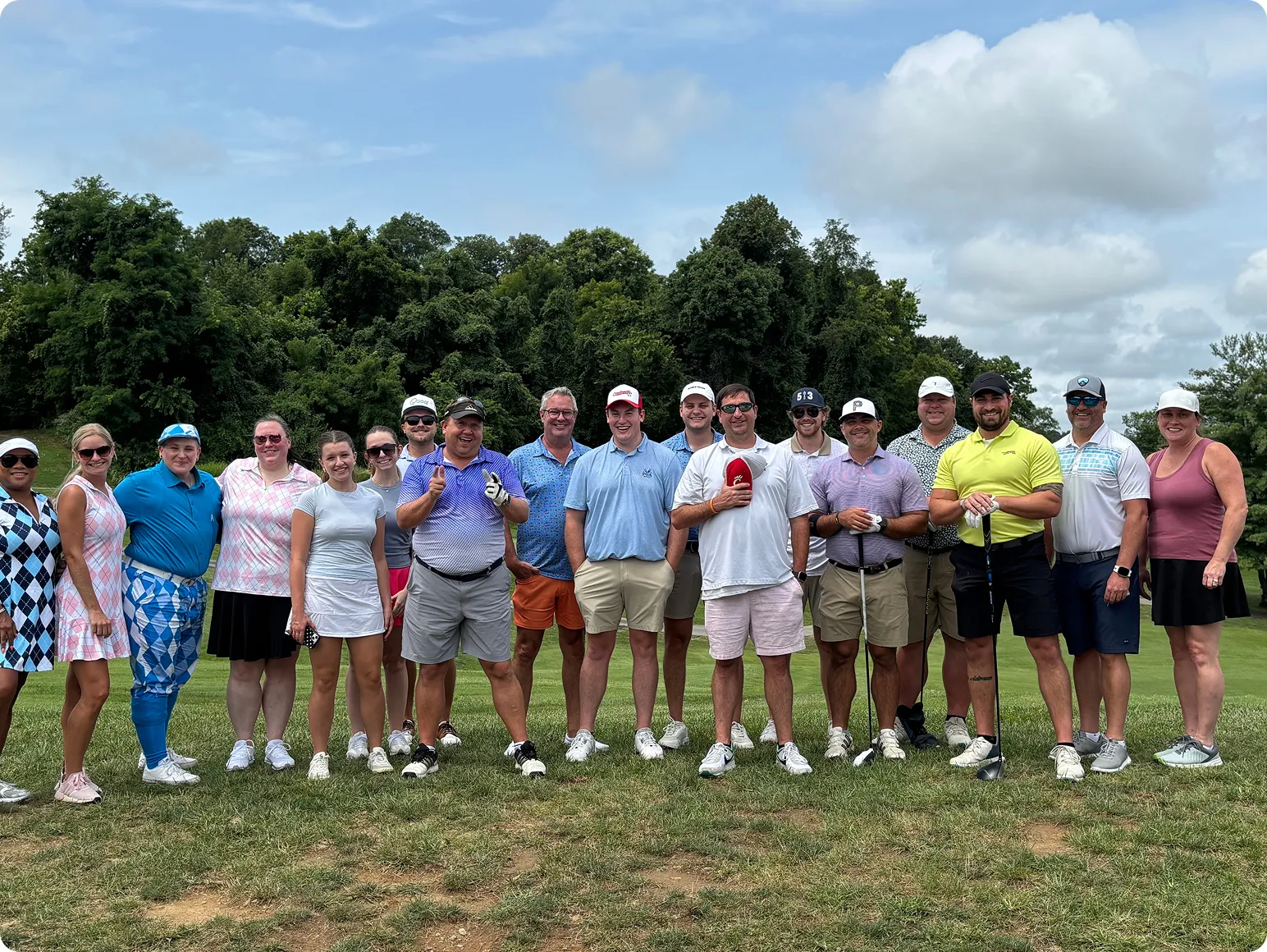 A team photo on a golf course, dressed in casual golf attire with trees and partly cloudy sky in the background.