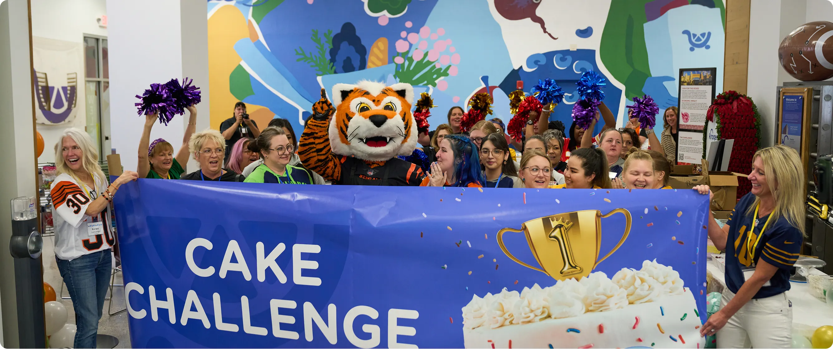 Group of people, including a person in a tiger mascot costume, holding a blue banner that reads 'Cake Challenge' with a gold trophy and cake image.