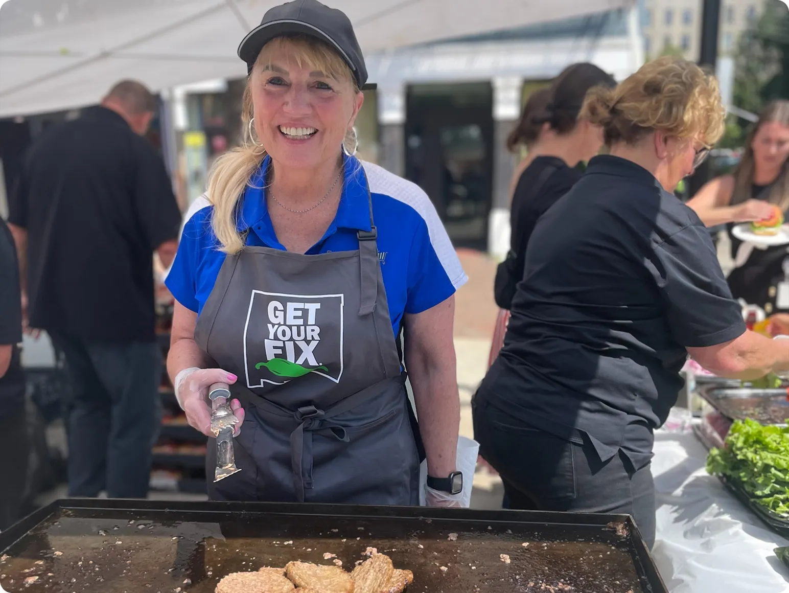 Smiling woman wearing a black cap and gray apron with 'GET YOUR FIX' text, cooking on a large flat grill outdoors.