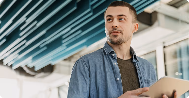 Young man in a denim shirt holding a tablet and looking to the side inside a modern building.