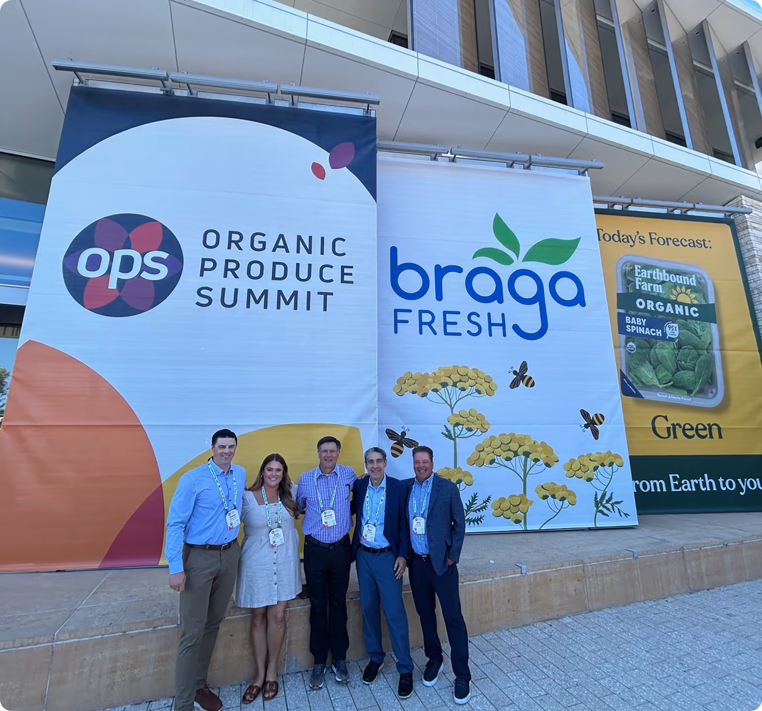 Five people standing and smiling in front of banners for Organic Produce Summit, braga Fresh, and Earthbound Farm Organic baby spinach.