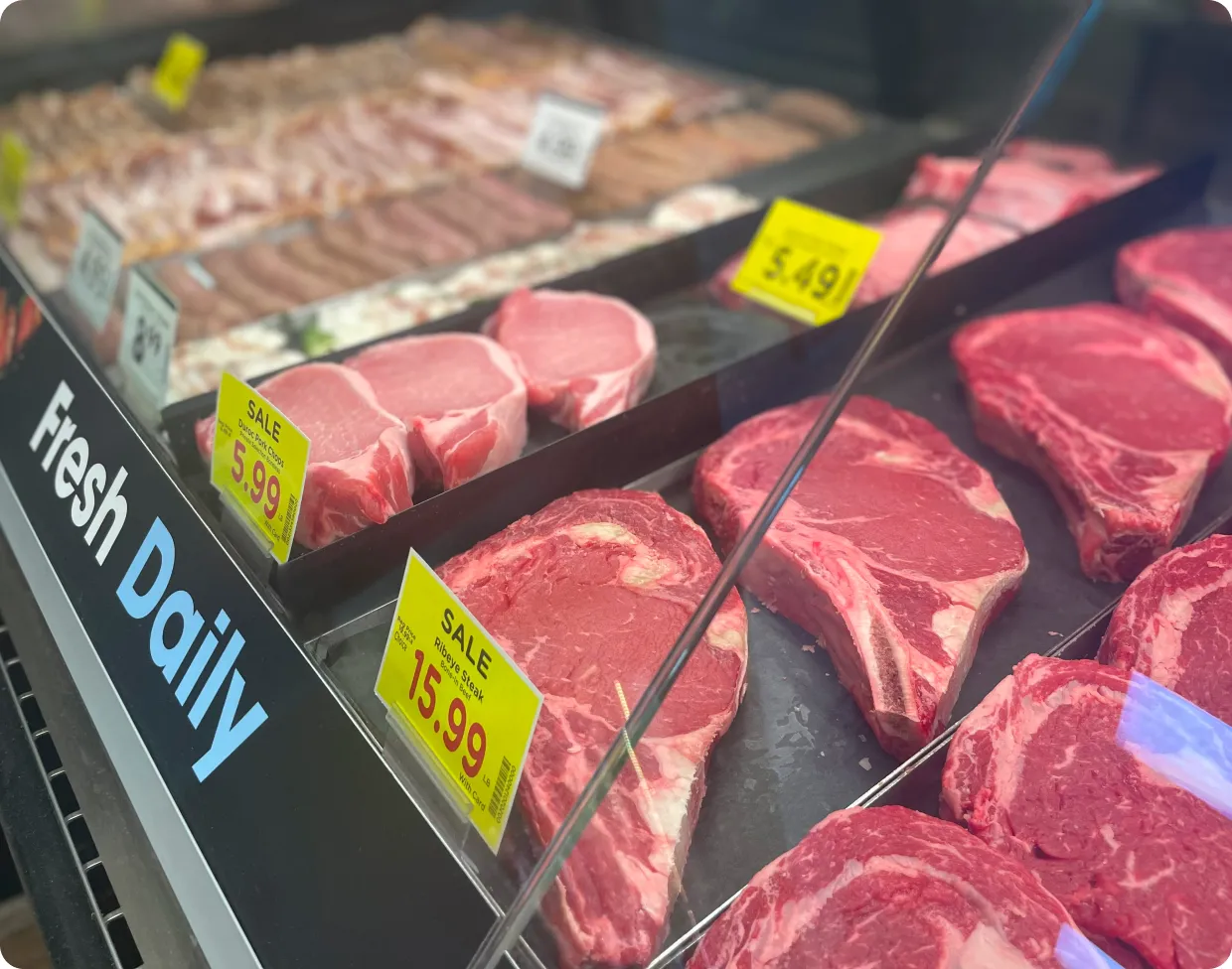 Display case of fresh raw meat cuts including pork chops and ribeye steaks with yellow sale price tags.