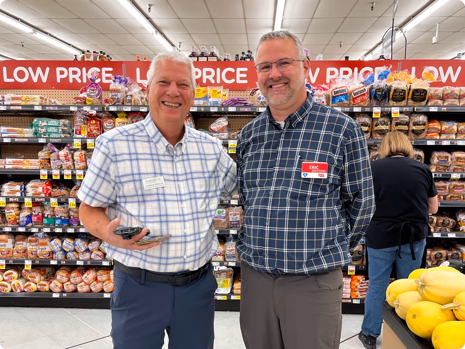 Two men smiling and standing in front of bread shelves in a grocery store, one holding a phone and the other wearing glasses.