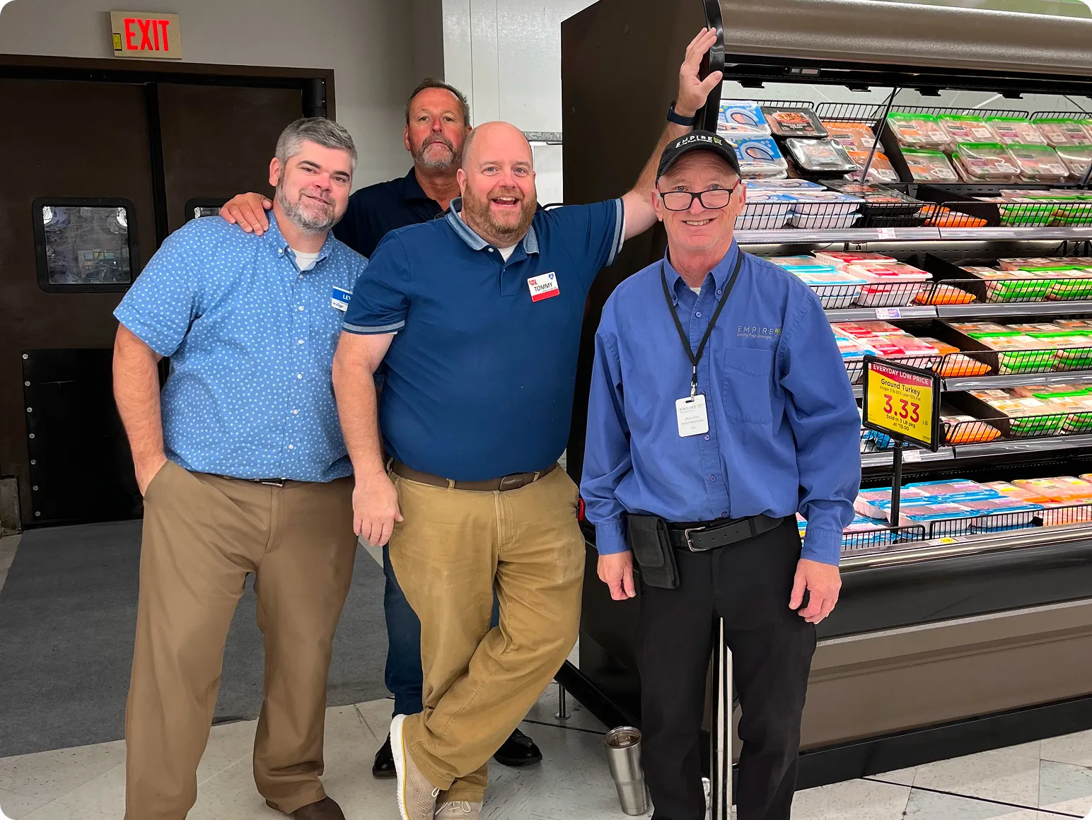 Four men posing inside a store next to a refrigerated meat display case with packaged grocery items.