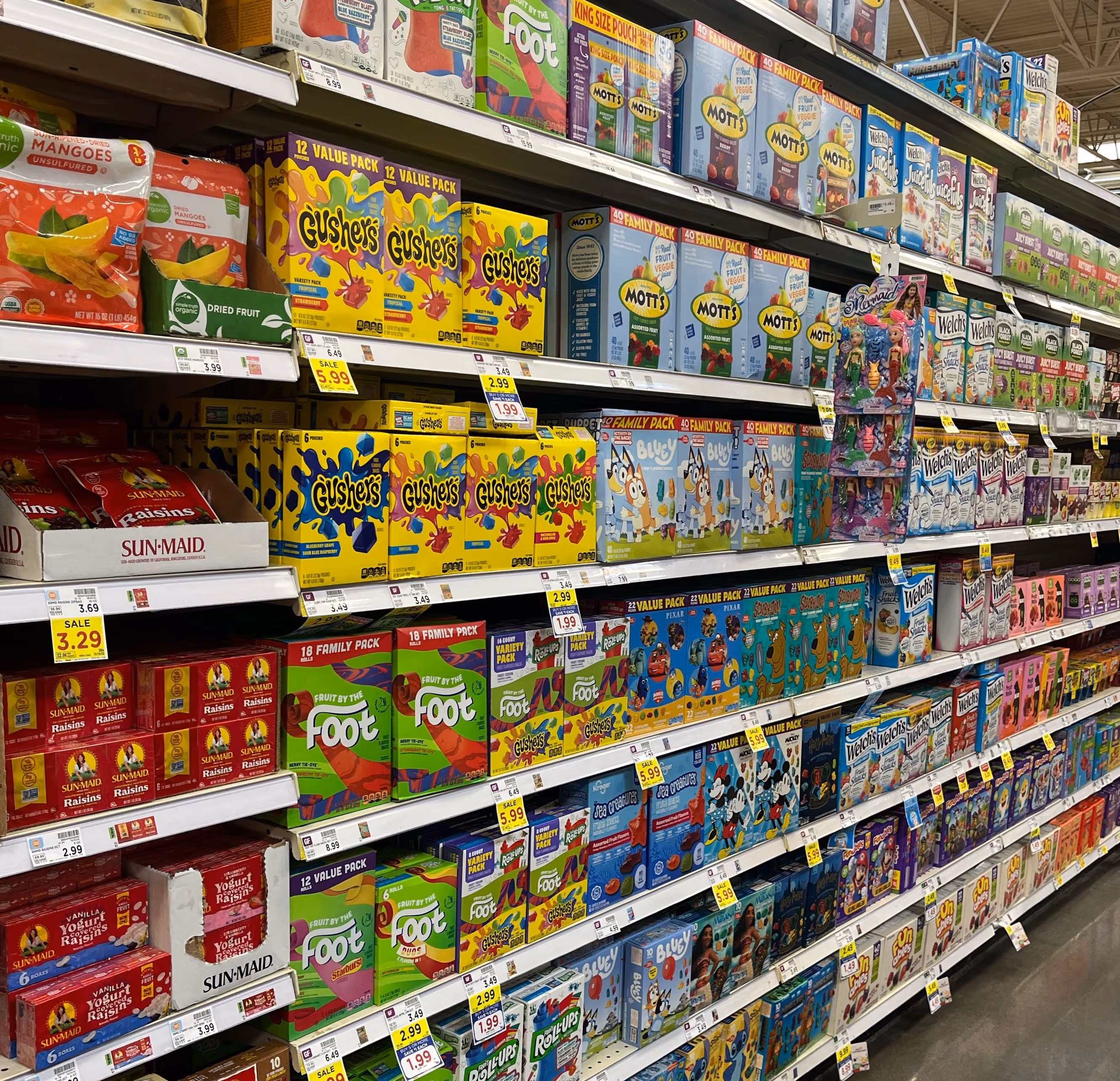 Grocery store aisle shelves stocked with various fruit snack brands including Gushers, Mott's, Welch's, Sun-Maid raisins, and Fruit by the Foot.